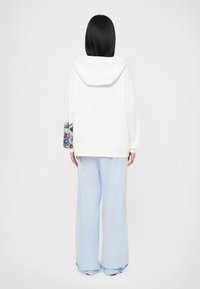 Persoană cu păr lung și negru, purtând un hanorac alb cu detalii colorate pe mâneci și pantaloni largi, cu dungi albastre deschise, stând cu spatele.