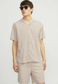 Ensemble de chemise à manches courtes à carreaux et de shorts en beige avec un motif de grille marron. Tissu léger, devant boutonné, coupe décontractée. Lunettes de soleil noires portées.