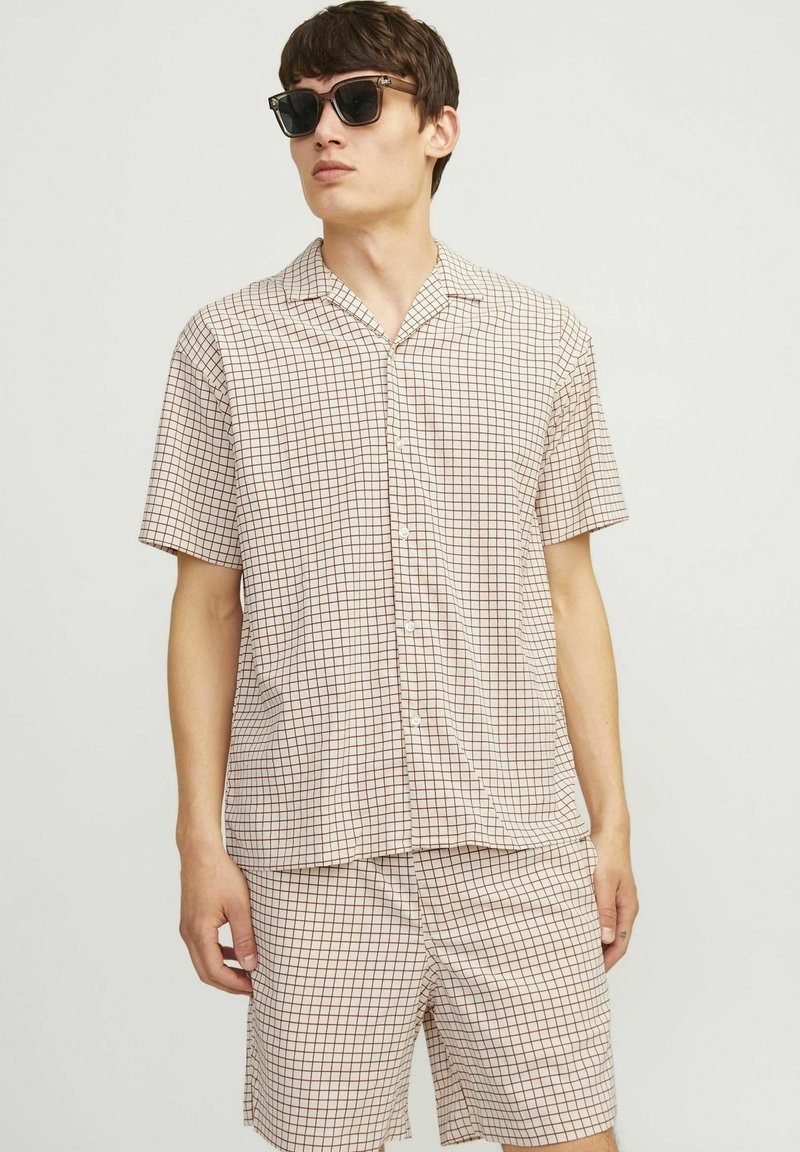 Ensemble de chemise à manches courtes à carreaux et de shorts en beige avec un motif de grille marron. Tissu léger, devant boutonné, coupe décontractée. Lunettes de soleil noires portées.