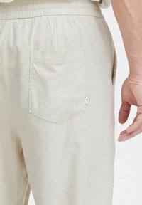 Close-up van beige lichte broek met elastische tailleband, achterzak en de rechterhand van een persoon met een zilveren armband zichtbaar.