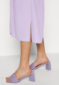 Chinelos roxos com um design de tiras trançadas e um salto quadrado grosso. O material parece suave com um acabamento brilhante.