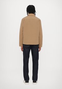 Braune gepolsterte Jacke mit Kragen, im geraden Schnitt und minimalistischem Design, kombiniert mit dunklen Jeans und weißen Sneakers.