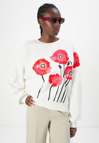 Felpa bianca con grandi fiori ricamati in rosso e rosa, dettagliati con steli neri. Il modello indossa occhiali da sole scuri e pantaloni beige chiaro.