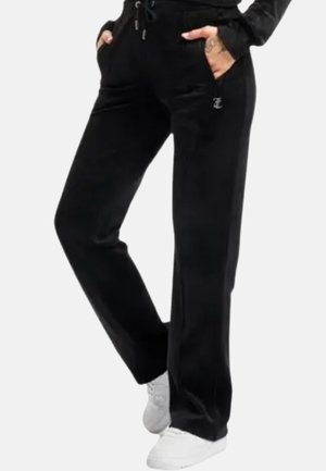 Persoon draagt zwarte velours wijde broek met zakken en witte sneakers, staat met handen in de zakken.