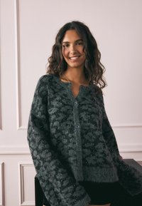 Sivih, prevelik kardigan s teksturiranim uzorkom, s velikim plavim gumbima i sjajnijim detaljima, napravljen od mekog, pahuljastog materijala.