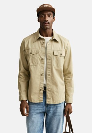 Mann trägt eine beige Hemdjacke über einem weißen Hemd, hellblaue Jeans, braune Mütze und hält den Griff einer braunen Tasche in der rechten Hand.