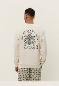 Felpa crema con grafica di palma nera e testo sul retro. Collo e polsini a coste. Abbinata a pantaloni a fantasia.