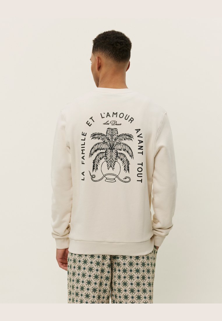 Sudadera crema con gráfico de palmera negra y texto en la parte posterior. Cuello y puños acanalados. Combinada con pantalones de estampado.