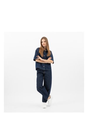 Giovane donna in piedi con le braccia incrociate, che indossa una camicia di jeans blu scuro, jeans e scarpe da ginnastica bianche su sfondo bianco.