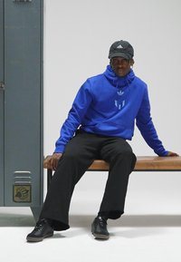 Camisola com capuz azul com um logótipo, calças pretas e ténis pretos. Sentado num banco de madeira ao lado de um cacifo cinzento. Design simples, visual casual.