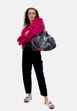 Femme en pantalon noir et sandales argentées, portant une veste texturée rose vif, tenant un grand sac à main sombre avec des patchs et des poignées colorées.