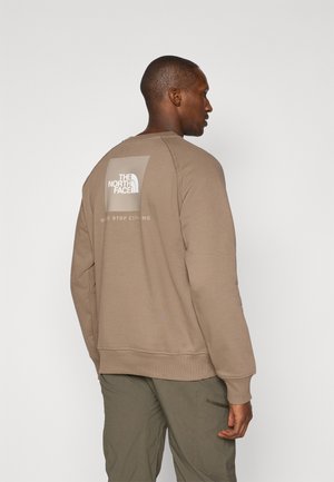 Brun sweatshirt med ribbede manchetter og nederkant, prydet med et stort hvidt logo på ryggen. Stoffet virker blødt med en glat overflade.