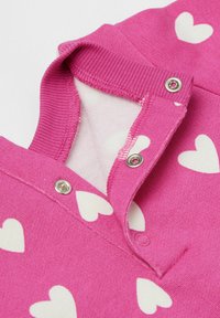 Rosa Sweatshirt mit weißen Herzmustern, ausgestattet mit einem gerippten Kragen und Druckknopfverschluss. Weiche Textur mit kontrastierender Innenauskleidung.