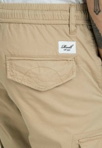Beige Cargo-Shorts aus Baumwollmischgewebe, mit elastischem Bund, einer Rückentasche mit Klappe und einem weißen Markenlabel.