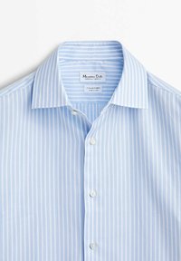 Chemise habillée bleu clair à rayures verticales blanches, col italien et boutons blancs, étalée à plat sur fond blanc.