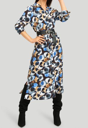 Femme portant une robe à manches longues boutonnée, à motifs de cercles bleus, noirs et beiges, cintrée avec une ceinture noire, associée à des bottes noires.