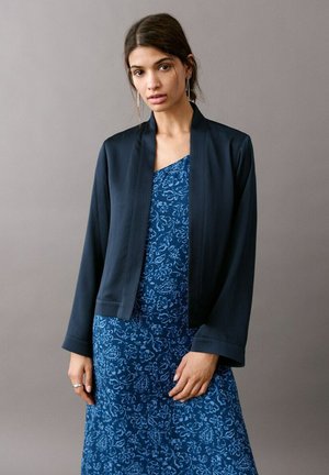 Femme portant une veste bleu marine foncé par-dessus une robe à motif floral bleu, debout devant un fond gris uni.
