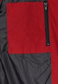 Czerwona łatka z widocznym przeszyciem na czarnej, gładkiej, podszytej powierzchni z zamykaną na suwak kieszenią po prawej stronie.