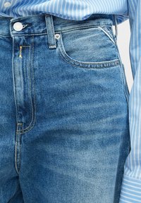 Artimas nuotrauka susidėvėjusių mėlynų džinsų su geltonais siūlais ir sidabriniu mygtuku, derinamų su mėlynos ir baltos juostelėmis marškiniais.