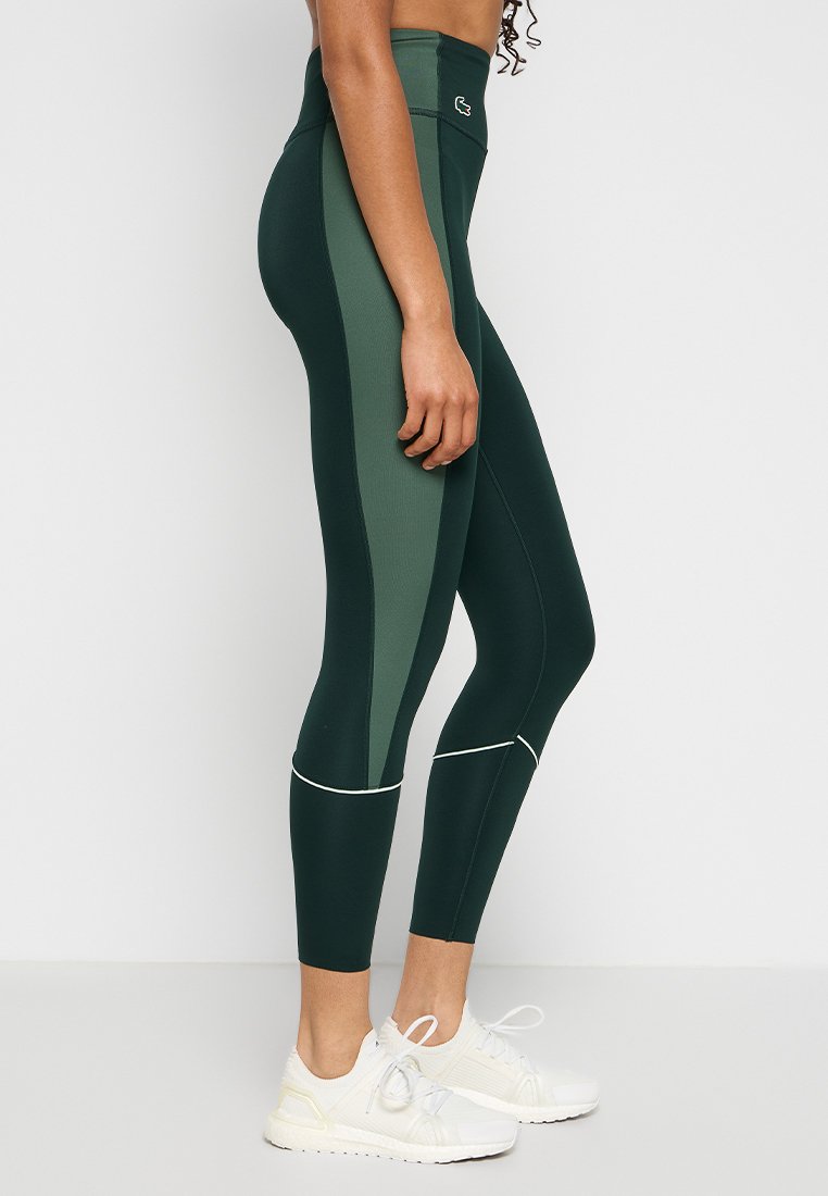 Leggings taille haute en vert foncé et panneaux vert clair, avec une texture lisse et des accents blancs. Présente un logo à la taille.