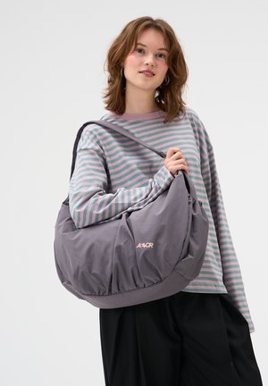 Junge Frau trägt ein gestreiftes Langarmshirt und schwarze Hose, hält eine große graue Umhängetasche mit AEVOR-Logo.