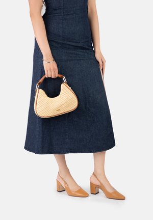 Woman in a dark denim midi dress holding a beige woven handbag and wearing tan block-heeled slingback shoes on a white background.