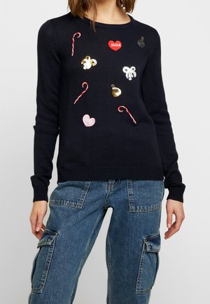 Vrouw die een marineblauwe trui draagt met kerstthema-patches, waaronder zuurstokken, harten, strikken en kerstballen, gecombineerd met blauwe cargobroek.