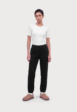 Persona joven de pie, vistiendo una camisa blanca de manga corta, pantalones jogger negros, calcetines blancos y zapatos marrones tipo slip-on con detalle de costuras.