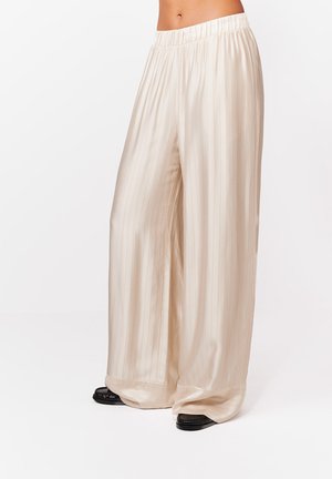Luźne kremowe spodnie z satyny z subtelnymi pionowymi paskami, z elastycznym pasem, noszone z czarnymi płaskimi butami.