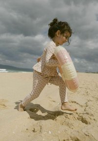 Ensemble de bain rose clair avec de petits motifs, comprenant un haut peplum et un legging ajusté. L'enfant tient une bouée gonflable pastel sur une plage de sable.