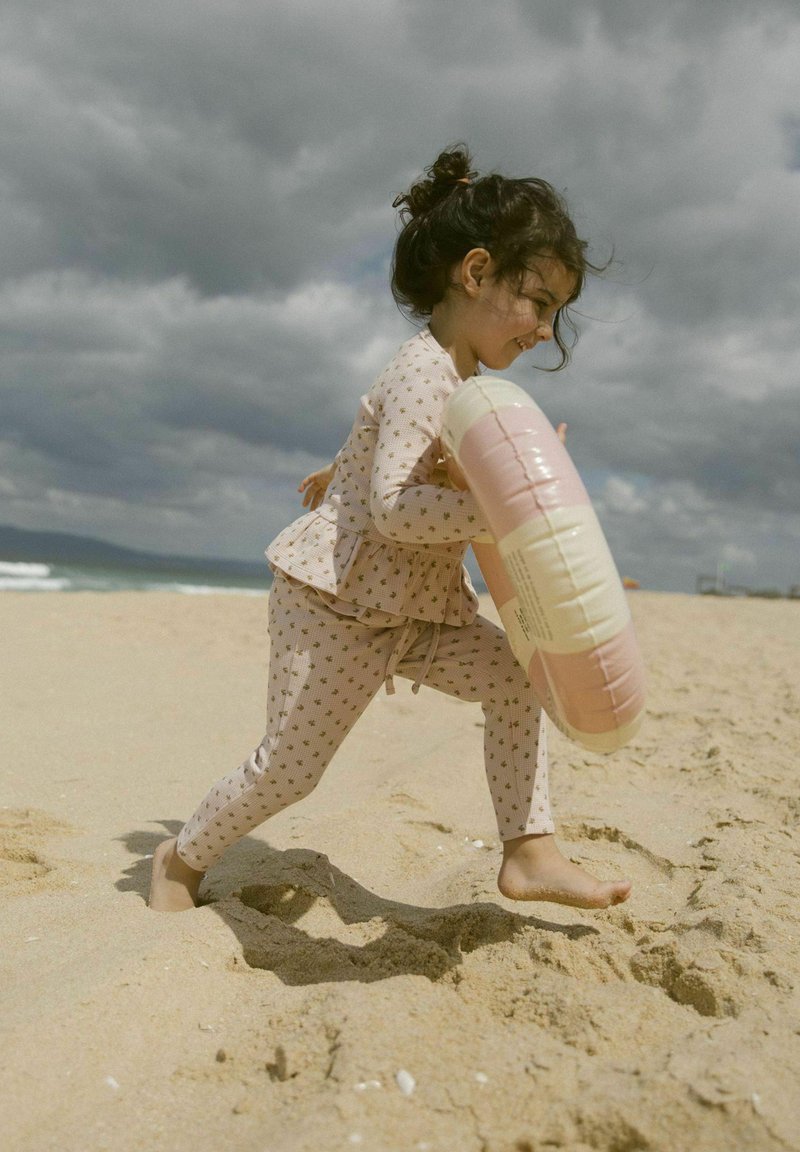 Ensemble de bain rose clair avec de petits motifs, comprenant un haut peplum et un legging ajusté. L'enfant tient une bouée gonflable pastel sur une plage de sable.