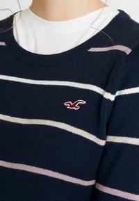 Sweter w granatowe paski z białymi i jasnoróżowymi poziomymi pasami. Ma okrągły dekolt i małe logo na piersi. Wykonany z dzianiny.