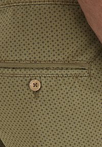 Buttoned back pocket on patterned olive green fabric, showing part of an arm resting nearby.