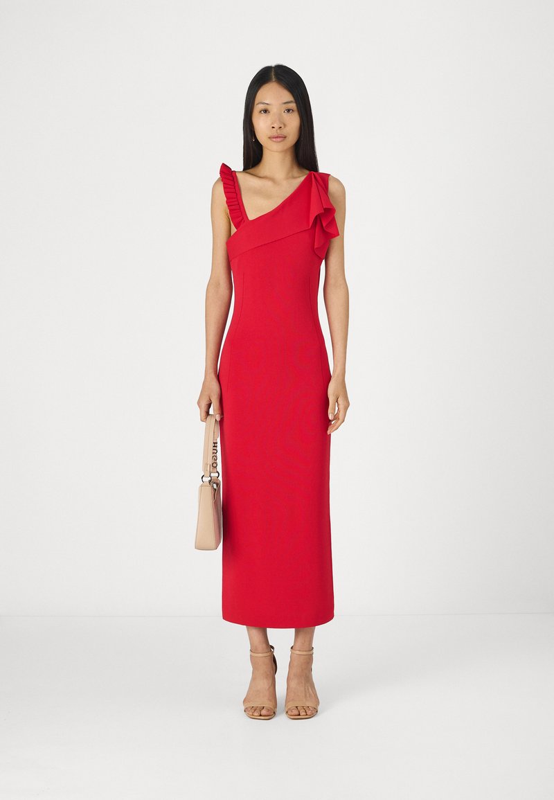 Czerwona sukienka na jedno ramię z falbaniastymi akcentami, dopasowany krój i długość do kolan. Modelka trzyma różową torebkę i ma na sobie cieliste sandały.