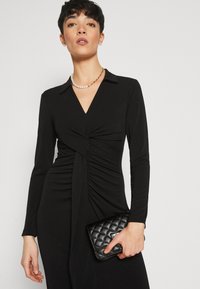Svart klänning med långa ärmar, V-ringning och snodd framsidesdesign. Tillsammans med en quiltad svart clutch och ett pärlhalsband.