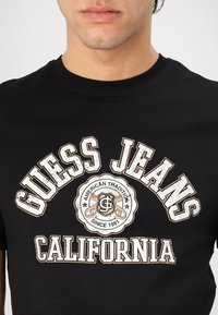 Camiseta de algodón negra con la inscripción "GUESS JEANS" en letras blancas y doradas, con un logo circular que dice "AMERICAN TRADITION SINCE 1981."