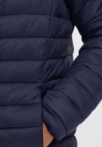 Közeli kép egy személyről, aki tengerészkék steppelt pufi dzsekit visel, kezét az oldalzsebben tartva.