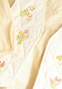 Quiltet tekstil med cremefarve, blomsterbroderi i pink, orange og grøn. Blødt stof med et struktureret, stribet mønster.