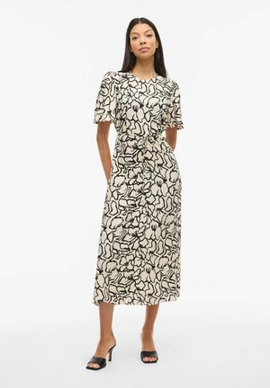 Femme debout portant une robe midi beige à imprimé floral abstrait noir, manches bouffantes, ceinture à nouer, et talons ouverts noirs sur fond blanc.