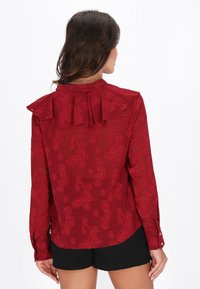 Blusa roja con estampado floral, escote con volantes, mangas largas y puños con botones. Tela semitransparente con detalles texturizados.