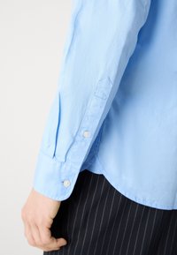 Personne portant une chemise bleu clair à manches longues avec poignet boutonné et un pantalon sombre à fines rayures, bras détendu sur le côté.