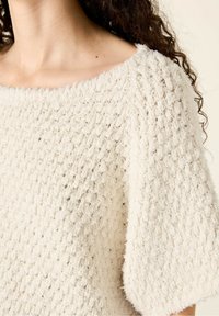 Top en tricot de couleur crème avec une finition texturée et duveteuse. Présente un décolleté large et des manches courtes, offrant une silhouette décontractée.