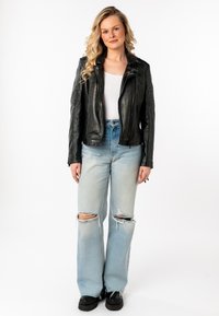 Schwarze Leder-Motorradjacke mit gerippten Schultern, kombiniert mit hellblauen, ausgebeulten Jeans im Weitformat und schwarzen Plateauschuhen.