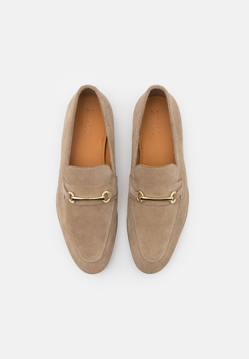 Mocassins en daim beige avec un accent chaîne en ton doré, présentant des détails cousus et un design à bout rond. Doublure intérieure lisse et semelle plate.
