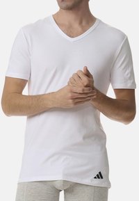 Vit kortärmad t-shirt med V-ringning i mjukt bomullsmaterial, med en svart Adidas-logotyp på nedre vänstra sidan. Enkelt design.