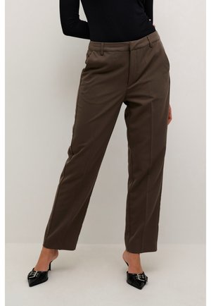 Vrouw draagt bruine op maat gemaakte broek en zwarte puntige muiltjes met hak en gespsluiting, staand tegen een effen achtergrond.