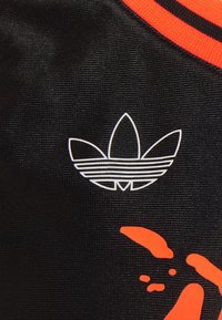 Svart tyg med en vit randig trefoillogotyp och en orange abstrakt design, vilket framhäver textur och kontrasterande färger.