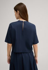 Marineblaues T-Shirt mit kurzem Arm, cropped Design und Schlüssellochdetail hinten, das mit einem Knopf gesichert ist. Glatte Textur, leichtes Material.