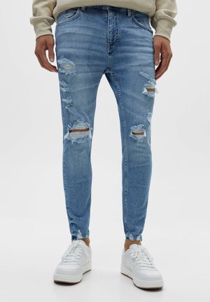 Homme portant un jean skinny bleu clair déchiré et des baskets blanches, debout devant un fond blanc uni.