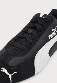 Gros plan d'une basket Puma noire avec des lacets noirs, logo Puma blanc sur l'avant et le côté, et un détail de bande blanche courbée sur le côté.